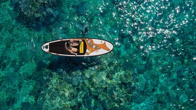 Eine Person paddelt auf einem Stand-Up-Paddle-Board über glitzerndes, klares Wasser, umgeben von bunten Korallen.