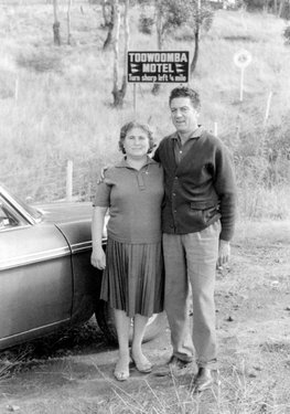 Die Frau trägt ein schlichtes Kleid und der Mann einen Pullover, beide posieren freundlich vor dem Toowoomba Motel-Schild.