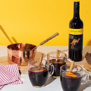 Ein kupferner Topf steht neben drei Gläsern mit heißem Getränk, dekoriert mit Zimtstangen und einer Flasche Rotwein.