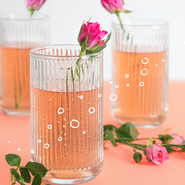 Drei Gläser mit rosafarbenem Getränk, dekoriert mit frischen Rosen und sprudelnden Blasen, auf einem hellen Untergrund.