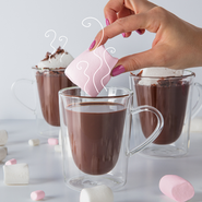 Eine Hand taucht einen großen, rosa Marshmallow in eine Tasse heiße Schokolade, umgeben von weiteren Süßigkeiten.