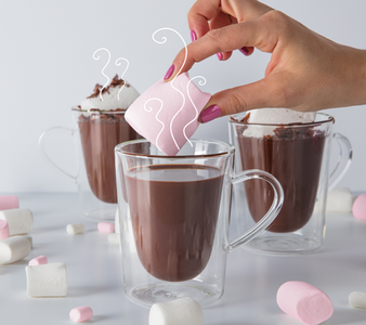 Eine Hand taucht einen großen, rosa Marshmallow in eine Tasse heiße Schokolade, umgeben von weiteren Süßigkeiten.