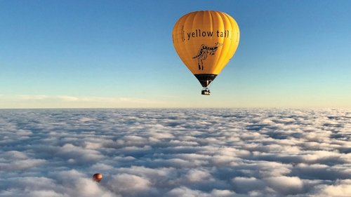 Ein leuchtend gelber Heißluftballon schwebt majestätisch über einer Wolkendecke, während die Sonne den Himmel erhellt.