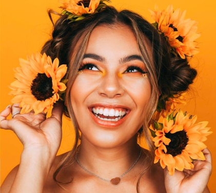 Eine fröhliche Frau mit Sonnenblumen im Haar lächelt strahlend vor einem leuchtend gelben Hintergrund.
