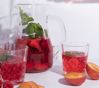 Ein Krug mit fruchtigem Getränk, gefüllt mit frischen Äpfeln und Minze, steht neben zwei Gläsern auf einem weißen Tisch.