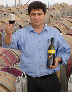 Ein Mann in einem blauen Hemd hält ein Glas Rotwein und eine Flasche Wein vor einer Reihe von Holzfässern.