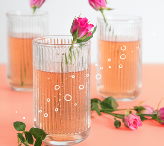 Drei Gläser mit rosafarbenem Getränk, dekoriert mit frischen Rosen und sprudelnden Blasen, auf einem hellen Untergrund.